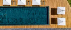 Aerial view of a women in a black swimsuit swimming in a rectangular pool, surrounded by sleek wooden decking and white parasols
