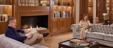 Wood-panelled room with a fireplace, bookshelves, and two people reading on grey chesterfield-style sofas