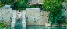 Woman in white walks down stone steps into a pool framed by tropical plants 