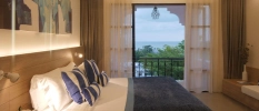 Hotel room with white bedding and walls, a navy throw and blue and white cushions, a wooden headboard and a blue and white wall painting, plus balcony doors open onto a balcony with greenery and sea views