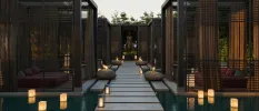 Stone walkway lined with lanterns with a Buddha statue at one end, and black cabin-style area with red daybeds