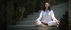 Woman in white sits cross legged with her eyes closed and palms up on a stone staircase