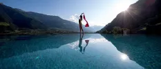 Woman in black swimsuit stands on the edge of a pool holding a red scarf, with the mountains and sun in the background