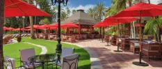 Wooden tables and chairs laid out next to grassy lawns and stone pathways, with red parasols lining the sides and tropical greenery surrounding them