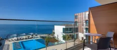 Balcony overlooking modern hotel building and a sea-facing swimming pool, set in a terrace with loungers