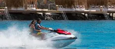 Couple on a jet ski driving past thatched-roof cabanas on the shore