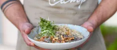 Man in a cream apron holds a white bowl with nutritious looking food topped with salad