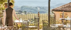 Waitress in a dark waistcoat setting the table on an outdoor terrace with black iron chairs and a view of the Tuscan hills