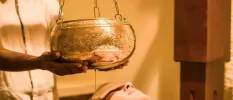 Woman lying down with an eye mask on as a therapist pours oil over her head from a golden bowl