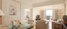 Hotel room living and dining area, with a glass dining table, bucket with a bottle and a fruit platter, next to a neutral sofa area and a balcony with a sea view