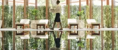 Staff member in dark shorts and a white shirt carries white towels through a room with an indoor pool, ivory-cushioned loungers and floor-to-ceiling windows showcasing tropical greenery