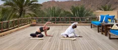 A woman in active wear with a staff instructor in traditional white dress, practicing yoga on a wooden deck surrounded by palm trees and mountains