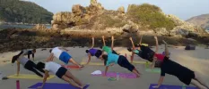 Group in active wear practice yoga on colourful yoga mats next to rocks under a blue sky