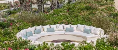 Circular ivory sofa with white and blue cushions fringed by bushes and flowers