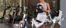 Woman in white shorts and a grey gym top holds a piece of cutting-edge equipment in a gym filled with exercise bikes