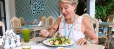 Woman in white tucks into a green salad with a glass of green juice next to her, and a sign that reads 'LOVE, LIVE, EAT' behind her