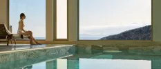 Woman in a printed swimsuit sits by an indoor pool gazing out large windows towards the sea and hillsides