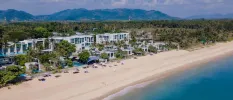 High-up view of white villas and pools on a white sandy beach, fronted by turquoise waves and backed by tropical greenery and mountains
