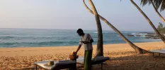 Waiter in traditional dress serving drinks to a table next to the ocean, with loungers and a hat left on one of them