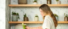 Woman in a white knitted shirt sits with a green smoothie with a kiwi garnish in a room with wooden shelves full of potted green plants