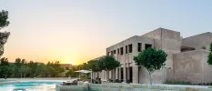 Greenery surrounds a swimming pool under a darkening sky, in front of a sand-coloured building with pillars