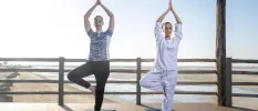 Two women in loose clothing in a yoga pose on a mat on a sunny terrace next to a sandy beach