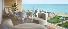 Glass-fronted balcony overlooking gardens and the sea, with neutral chairs and loungers and a table with aperol spritz