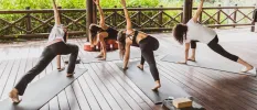 Four women in active wear practice yoga on mats on a wooden deck in an open-air pavilion