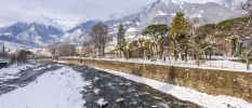 A frozen river under snowy peaks, with a tree-lined promenade and historic architecture