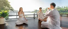 Three people in white sit cross-legged with their eyes closed holding their noses on a wooden deck next to a river
