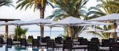 Cushioned wooden sun loungers in rows next to palm trees and under white umbrellas with the sea in the background
