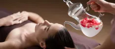Woman lies on her back with her eyes closed as a therapist pours liquid over her hair from a jug filled with rose petals