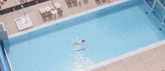 Woman in a white swimsuit floats on her back in a rectangular pool surrounded by wooden decking and loungers