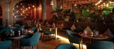 Glamorous restaurant with glossy plants, brown sofa-like seating and blue chairs, dark-wood tables and lantern-like lights