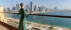 Woman in a green sheer dress looks out over a glass balcony at a white sandy beach, the sea and the Dubai skyline