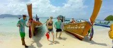 Guests exiting two longtail boats moored on a white sandy beach under a blue sky