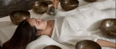 Woman in white lying down with her eyes closed, surrounded by sound bowls as a therapist uses a bell in front of her