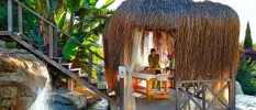 Woman lies on a bed in a thatched-roof open-sided cabana enjoying a massage in the tropical outdoors
