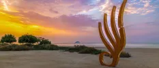 Wave-like sculpture on a sandy beach at sunset, with trees in the background