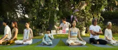 Group of adults and a child sit cross legged with their eyes closed on yoga mats in a tropical garden