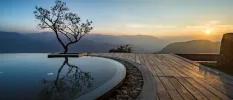 Curved edge of an infinity pool set in wooden decking with a tree next to it and views of misty mountains at sunset