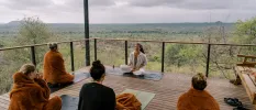 Group on yoga mats in brown blankets sit cross legged on a wooden deck overlooking the African wilderness