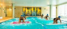 Group in black swimwear and blue swim hats balance on boards in an indoor pool during a group aqua fitness class