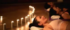 Woman lie on their backs with eyes closed on mats in a dark room lit by candles