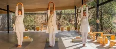 Three women in white, loose fitting clothing stretch their arms skywards in an open-sided pavilion surrounded by forest greenery