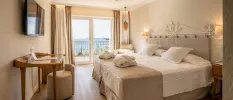 Neutral room with white bedding and cream throw with bathrobes on top, a dressing table and wall-hung TV, and full-length balcony with ocean views