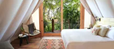 Airy room with inside drapes, white bedding, an aztec-style rug and wooden chair, and full-length balcony doors with a jungle view
