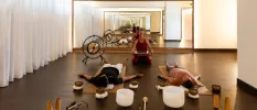 Two people lie on mats surrounded by singing bowls as a woman sitting next to a large gong heads up the class from the front