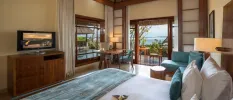 Bohemian-chic hotel room with wooden furnishings and white bedding, with blue cushions, a blue sofa and a blue throw, with a view outside to a furnished terrace and the ocean just beyond