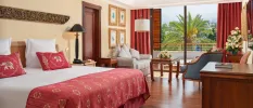 Hotel room with cream walls, white bedding, red soft furnishings, a blue chair and dark-wood furniture